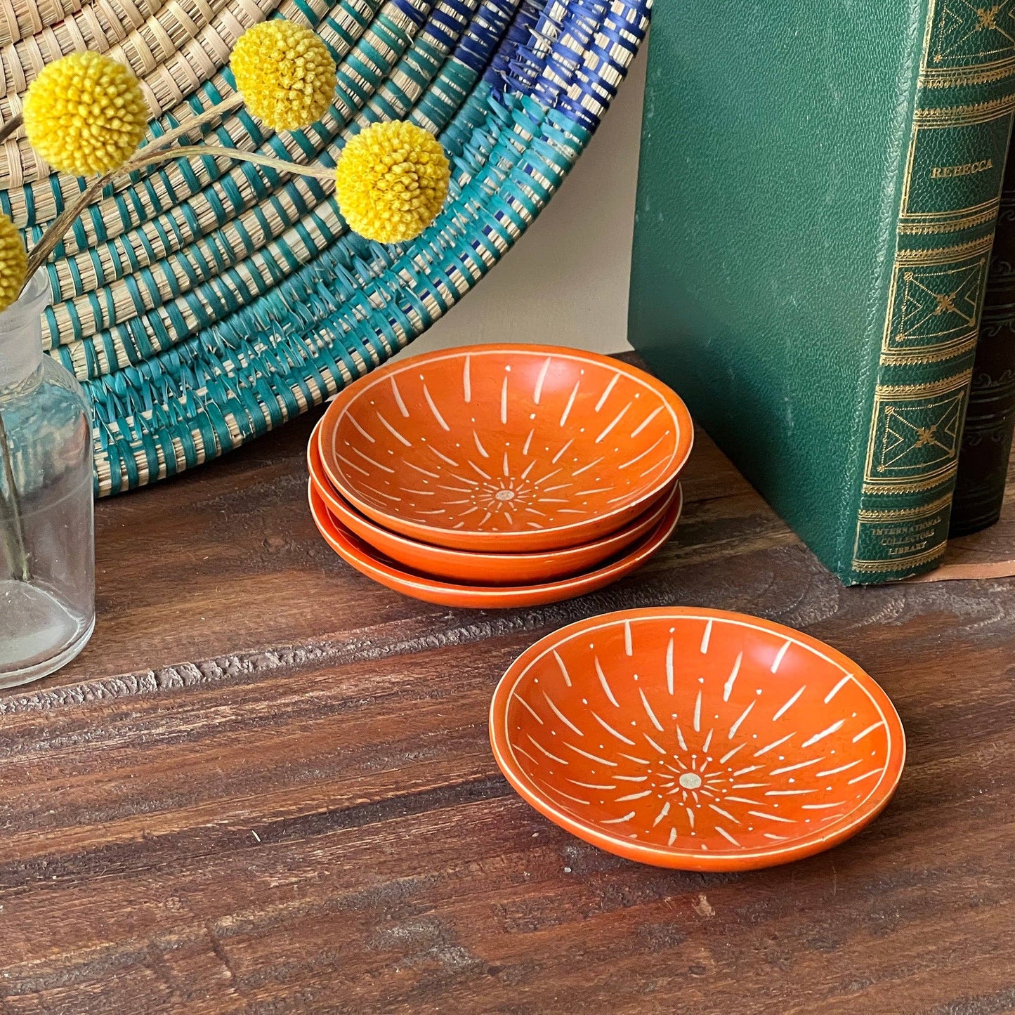 
                  
                    Stack of orange ceramic plates with decorative patterns on a wooden surface, with a book and decorative items in the background.
                  
                