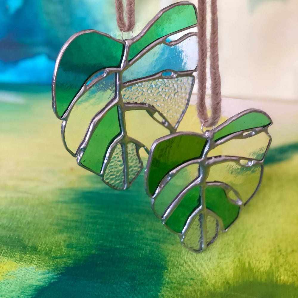 
                  
                    Two monstera leaf stained glass suncatcher with green, clear, and slightly iridescent panels and copper colored solder
                  
                